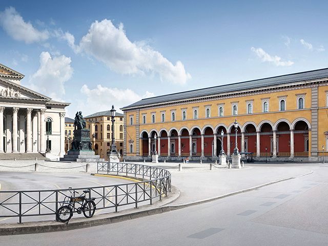 Historisches Gebäude mit Säulengang und Statue auf hellem Platz, Fahrrad abgestellt.